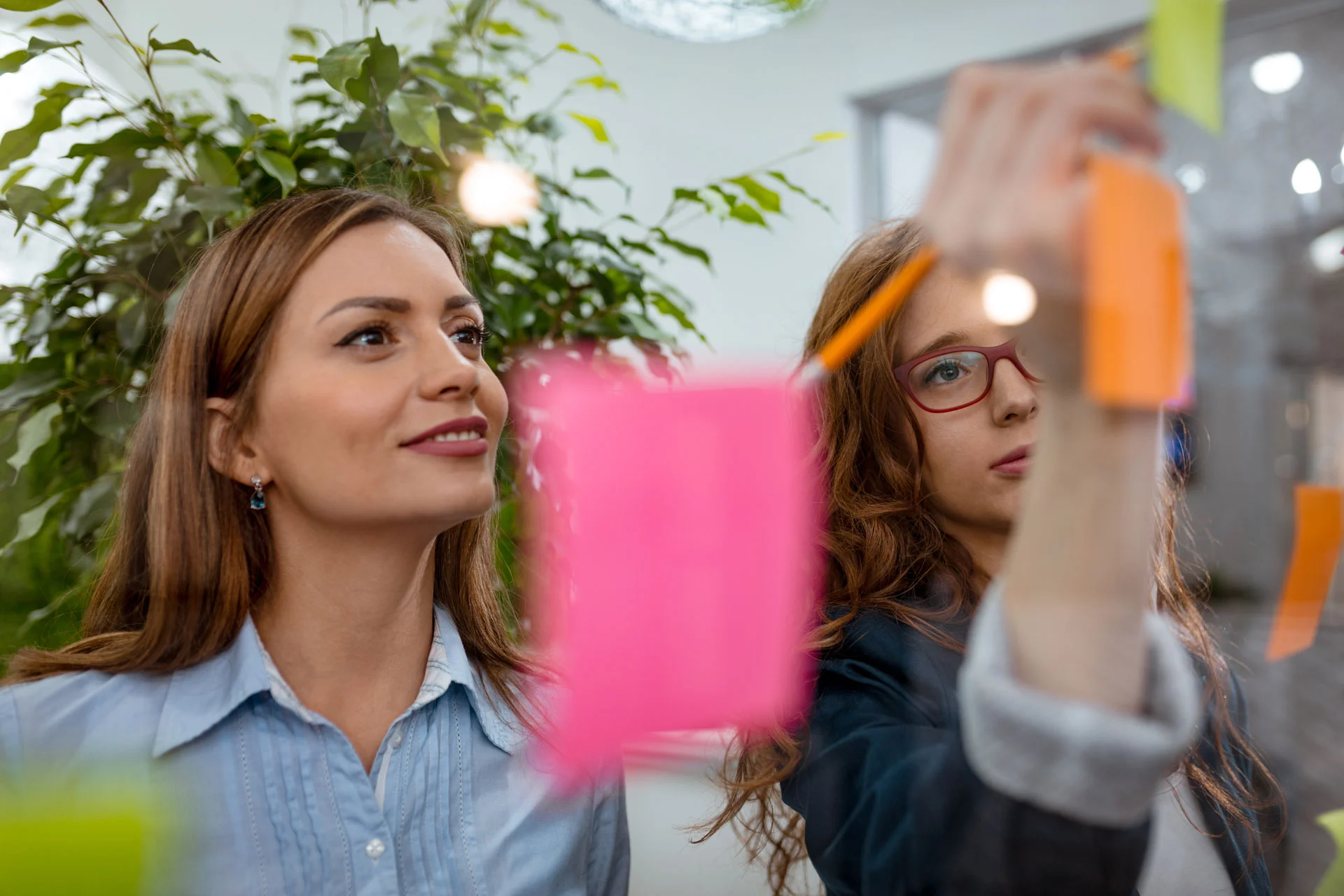creative-female-professionals-standing-discussing-office-glass-wall-with-sticky-notes-looking-post-it-note-wall